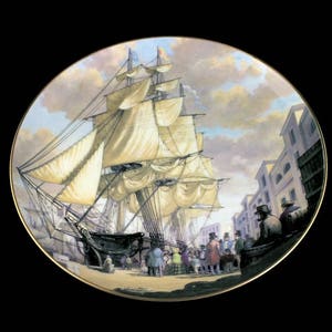May include: Decorative plate featuring a detailed painting of a tall ship docked in a harbor. The artwork depicts the ship with billowing sails, surrounded by buildings and figures in period clothing. The plate has a gold rim.