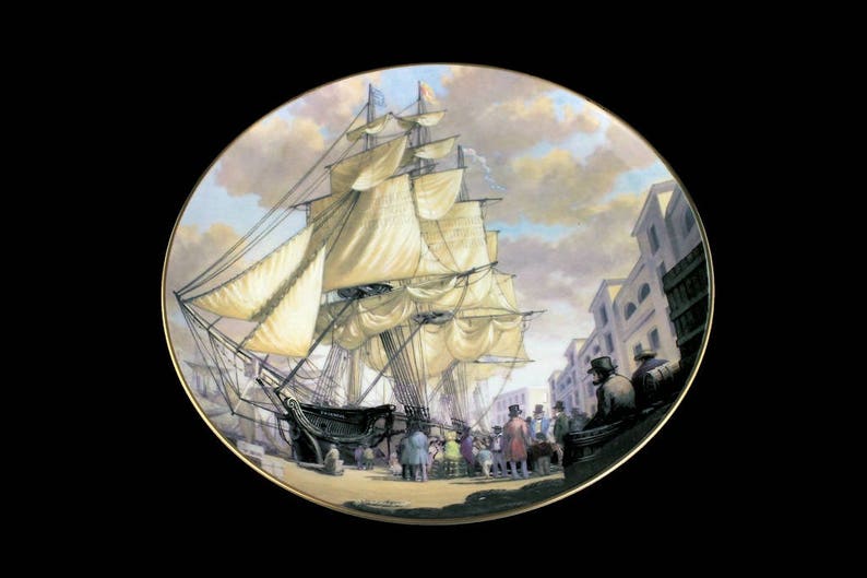 May include: Decorative plate featuring a detailed painting of a tall ship docked in a harbor. The artwork depicts the ship with billowing sails, surrounded by buildings and figures in period clothing. The plate has a gold rim.