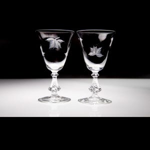 May include: Two clear wine glasses with etched leaf designs. The glasses have a detailed stem and base. The background is black and white, highlighting the delicate details of the glassware. Ideal for serving wine or other beverages.