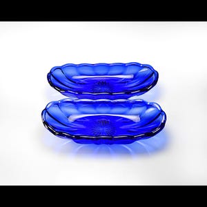 Puede incluir: Dos fuentes de vidrio azul cobalto. Cada plato ovalado tiene un borde festoneado y un diseño de estrella en el centro. Los platos se muestran sobre un fondo blanco.