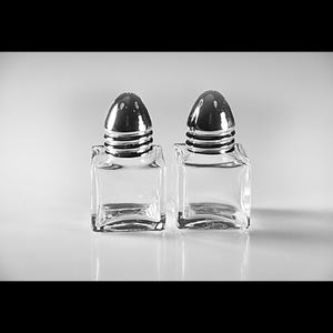 May include: Two clear glass salt and pepper shakers with silver tops. The shakers are square shaped and have a simple design.