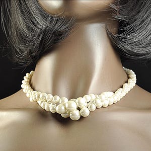 May include: Cream-coloured pearl necklace with a layered design. The necklace features multiple strands of small pearls and larger, prominent pearls at the centre. The necklace is displayed on a mannequin.