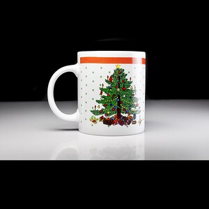 May include: White ceramic mug featuring a green Christmas tree design. The tree is adorned with red, yellow, and blue baubles and has a yellow star on top. The mug has a red and orange stripe around the top and a white background with green polka dots.