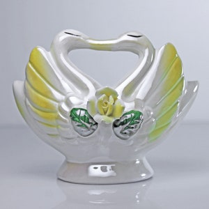 May include: White ceramic swan figurine with yellow and green accents. The swans are kissing and have a rose in the center of their necks.