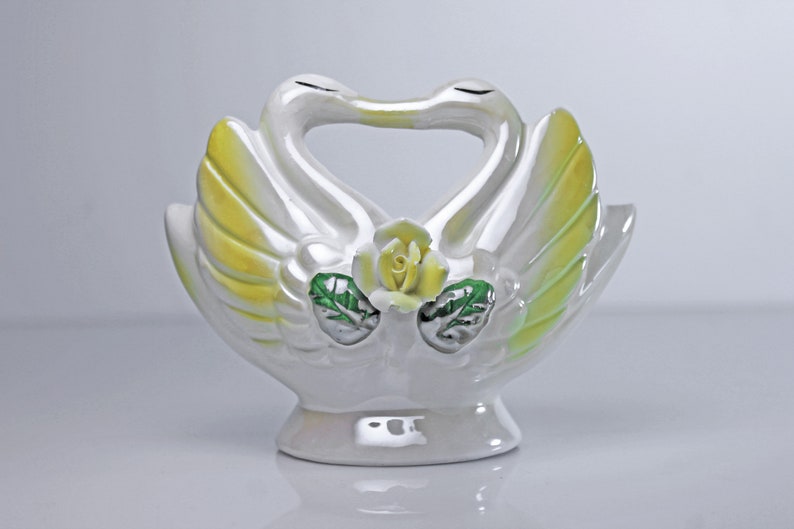 May include: White ceramic swan figurine with yellow and green accents. The swans are kissing and have a rose in the center of their necks.