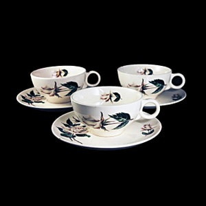 May include: Three white teacups with floral designs and matching saucers. The cups and saucers feature a white rose with green leaves.