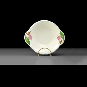 May include: Cream-coloured ceramic bowl with a scalloped rim, decorated with pink rose and green leaf motifs. The bowl is presented on a gold-toned metal stand, highlighting its floral design. A delightful item for serving or display.