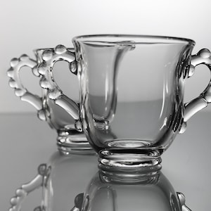 K&ouml;nnte beinhalten: Zwei klare Glasbecher mit perlenbesetzten Griffen. Die Becher sind leer und stehen auf einer reflektierenden Oberfl&auml;che.