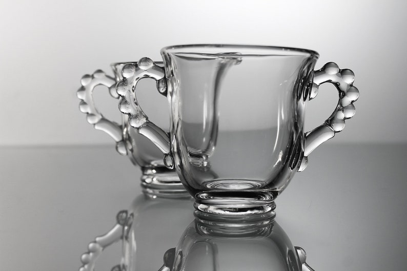 K&ouml;nnte beinhalten: Zwei klare Glasbecher mit perlenbesetzten Griffen. Die Becher sind leer und stehen auf einer reflektierenden Oberfl&auml;che.