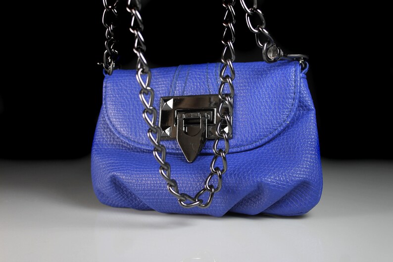 Pode incluir: Uma pequena bolsa azul cobalto com um acabamento texturizado. A bolsa apresenta uma al&ccedil;a de corrente prateada e um fecho de encaixe a condizer. O design &eacute; simples e elegante, adequado para v&aacute;rias ocasi&otilde;es.