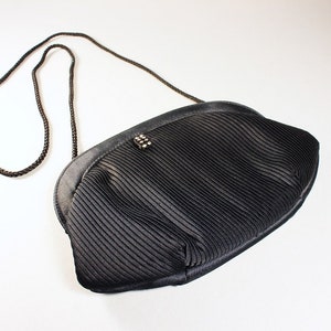 May include: A black pleated satin evening bag with a black chain strap. The bag has a small, decorative, silver-toned clasp.