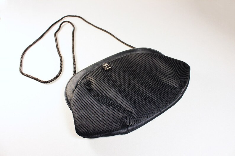 May include: A black pleated satin evening bag with a black chain strap. The bag has a small, decorative, silver-toned clasp.