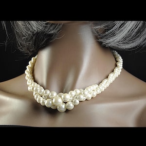 May include: A cream pearl necklace with a multi-strand design. The necklace features small pearls twisted together, with larger pearls clustered at the centre. The necklace is displayed on a mannequin.
