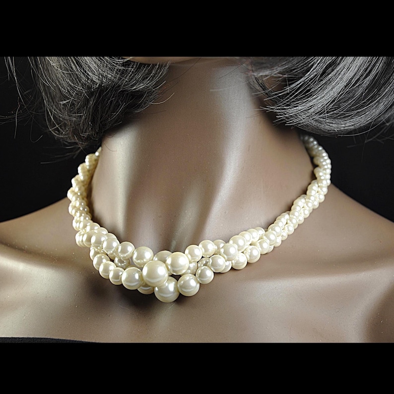 May include: A cream pearl necklace with a multi-strand design. The necklace features small pearls twisted together, with larger pearls clustered at the centre. The necklace is displayed on a mannequin.