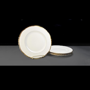 Puede incluir: Un juego de platos de porcelana blanca con borde dorado y borde festoneado. Los platos están apilados sobre una superficie gris sobre un fondo negro. Diseño clásico para cenas formales.