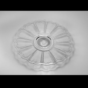Pu&ograve; includere: Un piatto da portata in vetro trasparente con un bordo ondulato e un motivo centrale di linee radianti. Il piatto presenta un motivo decorativo di linee curve attorno al bordo.