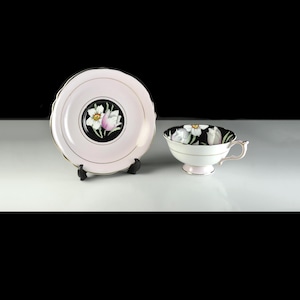 Puede incluir: Un juego de taza y platillo de té vintage. El platillo es de color rosa claro con un centro negro con un diseño floral. La taza es blanca con una base y asa rosas, y un interior negro con un estampado floral. Ribetes dorados adornan los bordes.