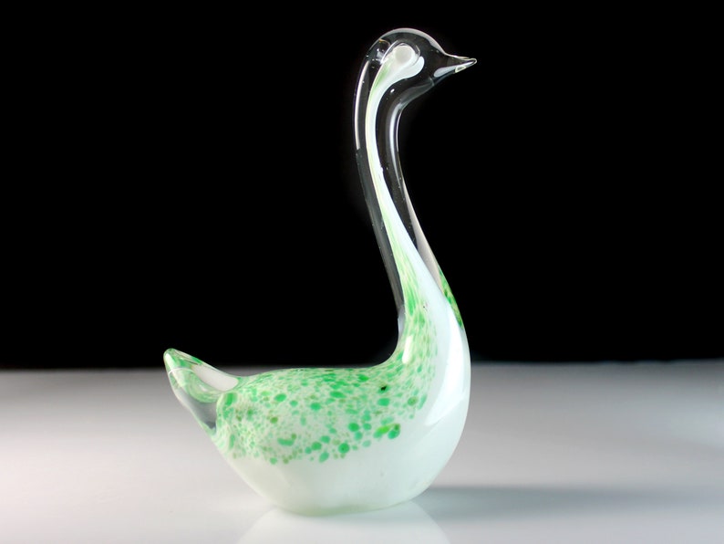 May include: A white glass swan figurine with green and white speckled accents. The swan has a long, curved neck and a small, round body.