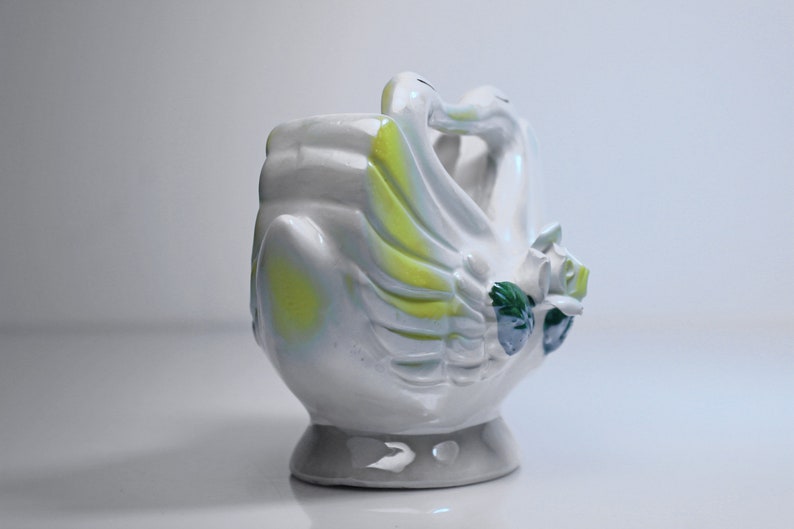 May include: White ceramic planter shaped like two swans with their necks intertwined. The swans have yellow and green accents and a floral design.