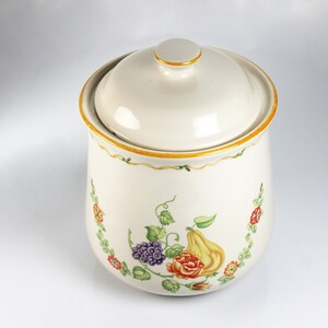 May include: A white ceramic canister with a lid, featuring a floral design with yellow, red, green, and purple accents. The canister has a gold trim around the top and bottom.