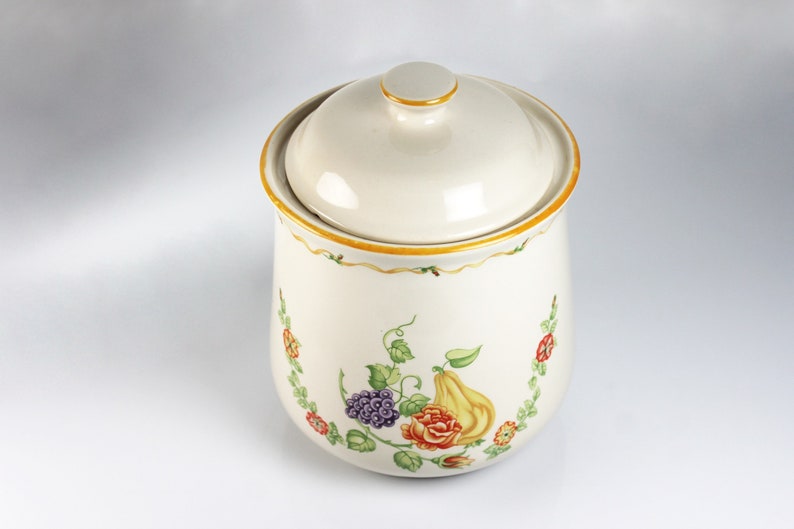 May include: A white ceramic canister with a lid, featuring a floral design with yellow, red, green, and purple accents. The canister has a gold trim around the top and bottom.