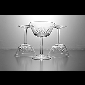 May include: Three clear glass champagne coupes with a cut glass design. The coupes are arranged in a row with the center coupe in focus.