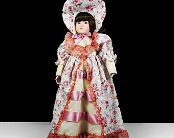 May include: A porcelain doll wearing a white dress with pink floral print and pink ribbons. The doll has brown hair and is wearing a white bonnet with pink floral print.