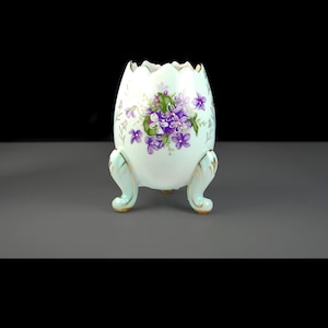 May include: A white porcelain egg-shaped planter with a scalloped rim, supported by three light blue feet with gold accents. The planter is decorated with a cluster of purple flowers and green leaves.