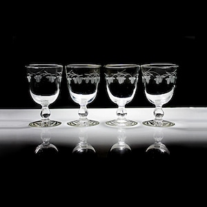May include: Four clear glass wine glasses with etched floral designs on the bowl. The glasses are arranged in a row on a white surface.