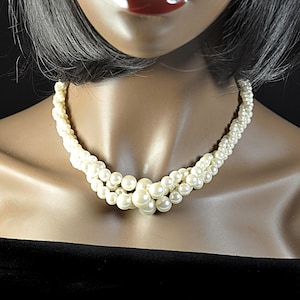 May include: Cream-coloured pearl necklace with a multi-strand design. The necklace features small pearls woven together, with larger pearls clustered at the centre. The necklace is displayed on a mannequin with a black top.