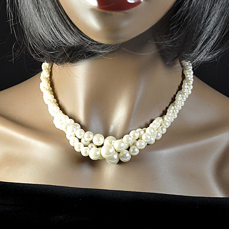 May include: Cream-coloured pearl necklace with a multi-strand design. The necklace features small pearls woven together, with larger pearls clustered at the centre. The necklace is displayed on a mannequin with a black top.