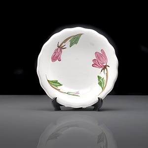 May include: White ceramic plate with a scalloped edge, decorated with pink flower and green leaf designs. The plate is displayed on a black stand against a dark background. The floral pattern adds a touch of elegance.