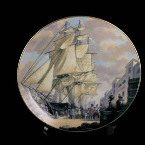 May include: Decorative plate featuring a detailed painting of a tall ship docked in a harbor. The artwork depicts a bustling scene with figures and buildings, set against a cloudy sky. The plate has a gold rim.