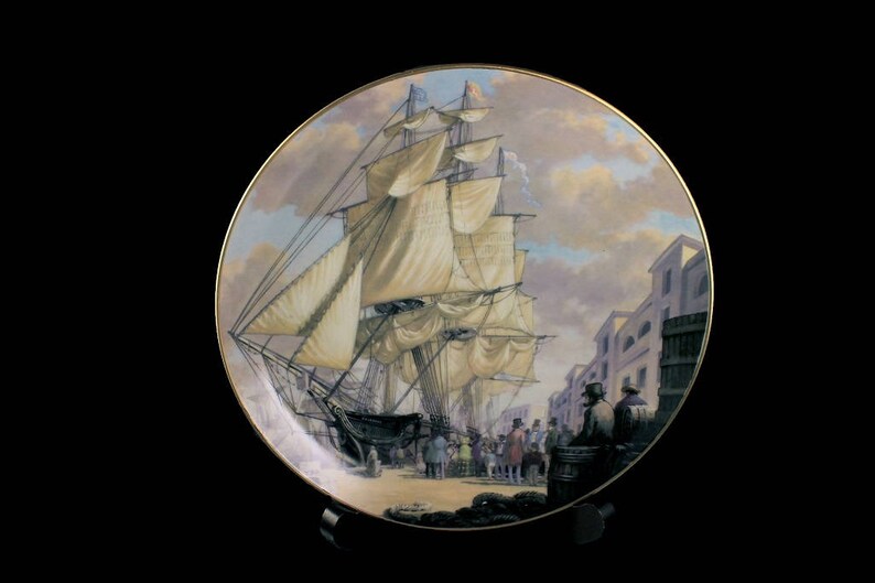 May include: Decorative plate featuring a detailed painting of a tall ship docked in a harbor. The artwork depicts a bustling scene with figures and buildings, set against a cloudy sky. The plate has a gold rim.