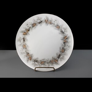 Op de afbeelding: Witte keramische dinerbord met een zwart en bruin dennenappelontwerp rond de rand.