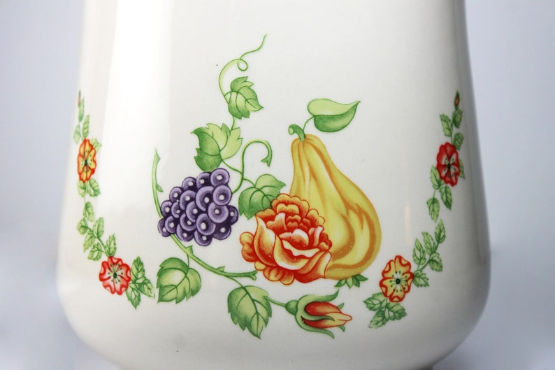 May include: White ceramic pitcher with a floral design featuring a pear, grapes, and a rose. The design is in shades of green, purple, orange, and yellow.
