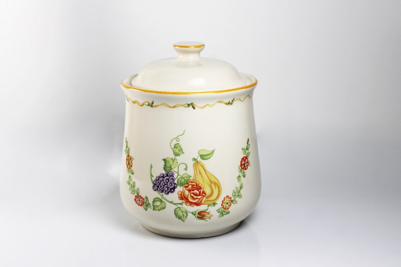 May include: White ceramic canister with a lid, featuring a floral design with a pear, grapes, and a rose. The design is in shades of green, yellow, red, purple, and brown.