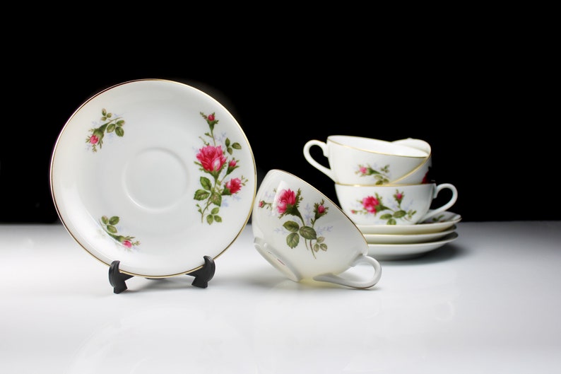 Peut inclure: Un ensemble de quatre tasses &agrave; th&eacute; et soucoupes blanches avec un motif floral repr&eacute;sentant des roses roses et des feuilles vertes. Les tasses et les soucoupes ont une bordure dor&eacute;e.