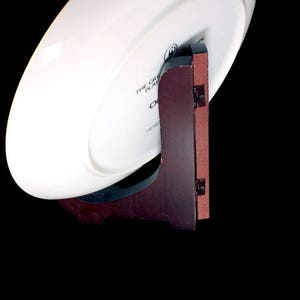 May include: A white oval plate displayed on a dark brown wooden plate stand. The stand has a reddish-brown backing and is attached with small black hinges. The plate has text that reads "THE GREAT PLATE".