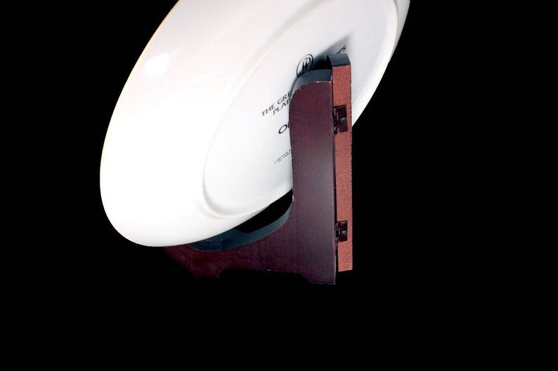 May include: A white oval plate displayed on a dark brown wooden plate stand. The stand has a reddish-brown backing and is attached with small black hinges. The plate has text that reads "THE GREAT PLATE".