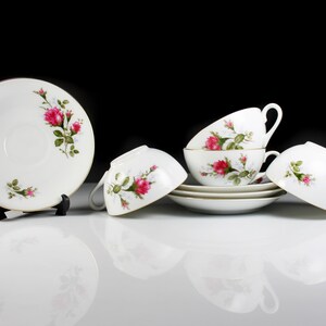 Peut inclure: Un ensemble de quatre tasses &agrave; th&eacute; et soucoupes blanches avec un motif floral compos&eacute; de roses roses et de feuilles vertes. Les tasses et les soucoupes sont empil&eacute;es les unes sur les autres.