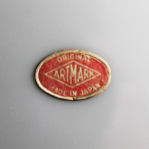 Puede incluir: Una pegatina ovalada roja con letras doradas que dice "ORIGINAL ARTMARK MADE IN JAPAN".
