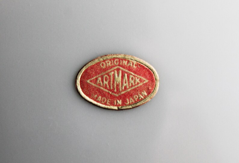 Puede incluir: Una pegatina ovalada roja con letras doradas que dice "ORIGINAL ARTMARK MADE IN JAPAN".