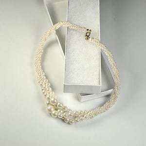 May include: A cream pearl necklace with a gold clasp, presented in an open white jewellery box. The necklace has multiple strands of varying pearl sizes, creating a layered effect. The pearls have a lustrous sheen.