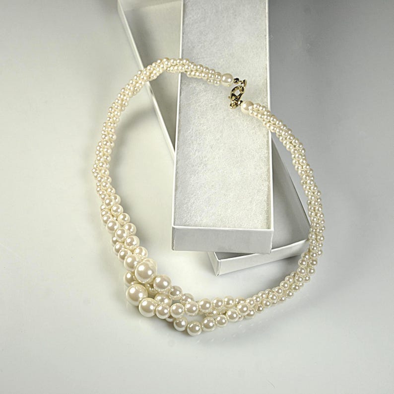 May include: A cream pearl necklace with a gold clasp, presented in an open white jewellery box. The necklace has multiple strands of varying pearl sizes, creating a layered effect. The pearls have a lustrous sheen.
