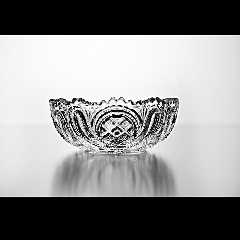 Pu&ograve; includere: Una ciotola di vetro trasparente con un bordo smerlato decorativo. La ciotola presenta motivi dettagliati e un motivo centrale a diamante. Si trova su una superficie riflettente, creando un effetto specchio. Il design &egrave; vintage ed elegante.