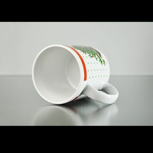 May include: White ceramic mug with a green and red Christmas tree design. The mug has a white handle and a red stripe around the top.