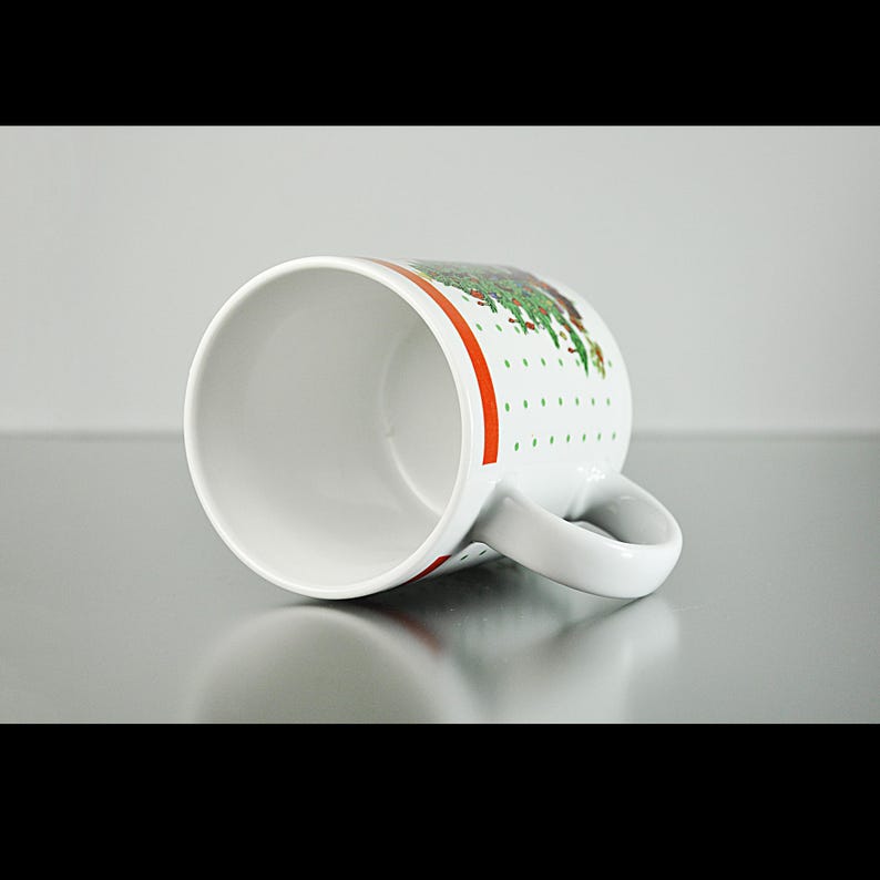 May include: White ceramic mug with a green and red Christmas tree design. The mug has a white handle and a red stripe around the top.