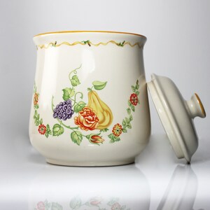 May include: White ceramic cookie jar with a floral design featuring a pear, grapes, and a rose. The lid is off and the jar has a gold trim.