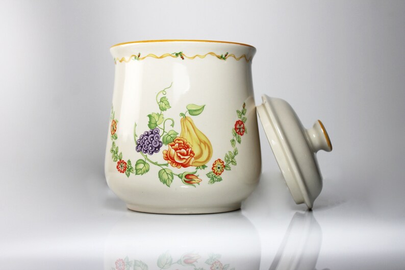 May include: White ceramic cookie jar with a floral design featuring a pear, grapes, and a rose. The lid is off and the jar has a gold trim.